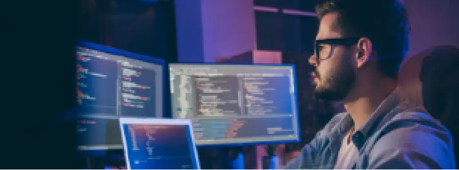 Man working at multiple computer screens displaying code, representing software development and cybersecurity monitoring.